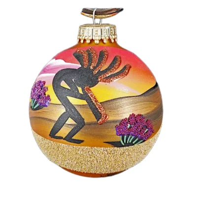 Hand-painted glass sphere ornament depicting a black dancing Kokopelli flute player with copper glitter accents, desert flora, and sunset background.