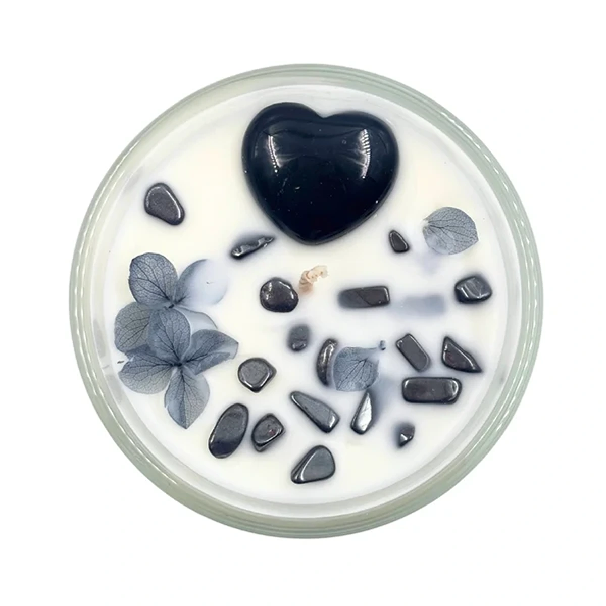 Round candle in a glass jar with white wax, featuring a large black heart centerpiece, scattered gray stones, and blue-gray leaves.